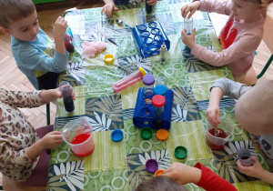 Dzieci siedzą przy stoliku i barwią wodę tuszem w różnych kolorach