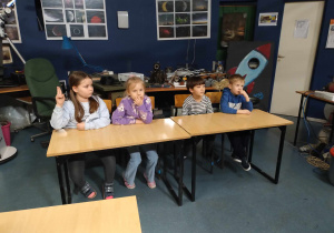 Dzieci siedzą w ławkach, obserwują eksperymenty, które wykonuje prowadzący.