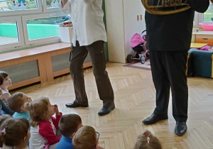 Dzieci siedzą na dywanie, panowie prezentują instrument