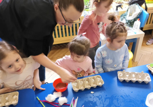 dziewczynki siedzą przy stoliku i wysiewają rzeżuchę, mam Zosi pomaga dzieciom w zadaniu