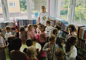 dzieci stoją w dziale książek dla dzieci, słuchają Pani bibliotekarki