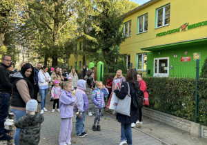 dzieci wraz z rodzicami stoją pod przedszkolem i słuchają od pani przewodnik informacji na temat zadań do wykonania i otrzymują mapki trasy
