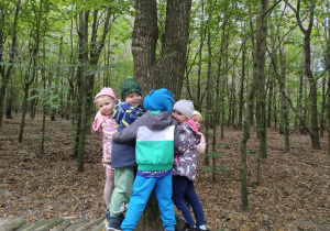 Dzieci przytulają się do drzewa stojąc na drewnianym mostku.