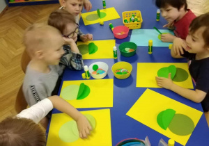 Dzieci z grupy zielonej siedzą przy stolikach i według instrukcji nauczyciela wykonują samodzielnie żabki.