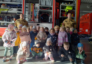 Dzieci z grupy zielonej wraz z dwoma strażakami ustawione koło wozu strażackiego do zdjęcia grupowego.