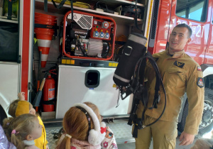 Strażak prezentuje dzieciom butle i maskę z tlenem, która Strażacy mają na plecach podczas akcji gaszenia pożaru.