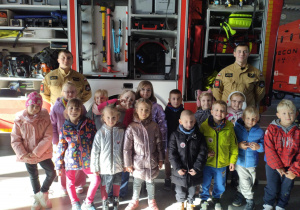 Dzieci z grupy srebrnej wraz z dwoma strażakami ustawione koło wozu strażackiego do zdjęcia grupowego.