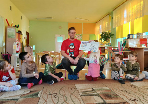 dzieci siedzą w kole na dywanie, tata Zuzi czyta książkę, a dziewczynka pokazuje ilustrację