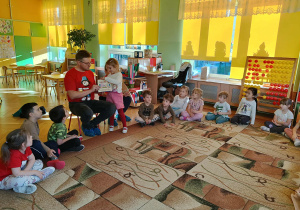 dzieci siedzą w kole na dywanie, tata Zuzi czyta książkę, a dziewczynka pokazuje ilustrację